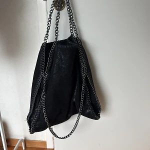 Kedjeväska - Cool svart axelväska i mockaimitation med glansig finish och kraftiga silverfärgade kedjor som axelremmar och längs kanterna. Väskan har en mjuk, slouchy form och dragkedja på insidan. Perfekt för dig som gillar edgy och trendig stil.