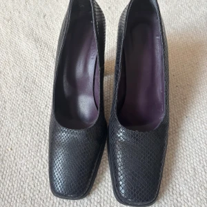 Svarta pumps i  skinn 39 - Snygga svarta pumps i äkta skinn med  fyrkantig tå. Klacken är bred och stabil, vilket ger en cool och modern siluett. Tillverkade i Italien och fodrade med mörklila insida. Perfekta för dig som gillar statementskor.