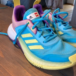 Adidas x LEGO sneakers blå/gul 36⅔ - Färgglada Adidas x LEGO sneakers i blått, gult och lila med coola LEGO-detaljer på hälen. Skorna har snörning, mesh- och syntetmaterial samt klassiska tre ränder i gult på sidan. Perfekta för dig som gillar unika och lekfulla sneakers.