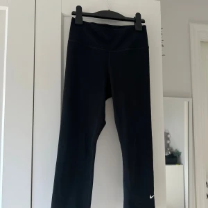 Svarta Nike Dri-Fit trekvarts leggings XS - Svarta trekvarts leggings från Nike i Dri-Fit material, perfekt för träning. Hög midja och tight passform, med klassisk vit Nike-logga vid benet. Snygga och bekväma tights som sitter på plats under hela passet.