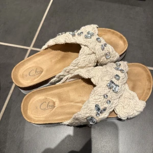 Beige sandaler med flätad textil och stenar - Snygga sandaler från Clou med breda, flätade band i beige textil och glittrande stenar på ovandelen. Skorna har en platt sula med korkdetalj och vit undersida. Perfekta för sommarens alla tillfällen när du vill ha något både bekvämt och trendigt.