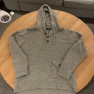 Grå stickad hoodie från French Connection - Mysig grå stickad hoodie från French Connection med huva och knappar framtill. Tröjan har en praktisk ficka med knapp på magen och ribbade muddar vid ärmslut och nederkant. Perfekt för kyliga dagar och enkel att matcha med jeans. Är i storlek L men passar bra som M