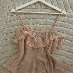 Beige volangtop med spetsdetaljer - Supersöt beige linnetopp med tunna axelband, volanger och broderade spetsdetaljer. Toppen har en romantisk känsla med blommig spets framtill och volangkant runt hela överdelen. Perfekt för dig som gillar en feminin och luftig stil.