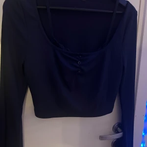 Mörkblå croppad långärmad topp - Säljer en mörkblå croppad topp med långa ärmar och ribbad struktur. Toppen har en snygg, djup rundad urringning framtill och tre små knappar som detalj. Perfekt att styla med höga jeans eller kjol för en trendig look.