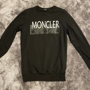 Svart Moncler sweatshirt  - Svart sweatshirt från Moncler med stor vit logga framtill och diskret svart tryck under. Tröjan har rund hals, långa ärmar och ribbade muddar vid ärmslut och nederkant. Tillverkad i mjuk bomull för en skön känsla.