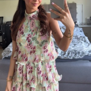 Blommig volangklänning med hög krage - Supersöt blommig klänning med volanger i lager och hög krage. Klänningen har pastelliga toner i rosa, grön och vit, och är ärmlös med en flowy passform. Perfekt för sommarens alla tillfällen!