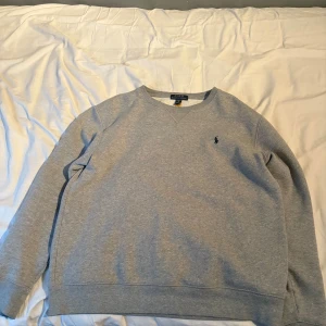 Grå sweatshirt från Polo Ralph Lauren - Klassisk grå sweatshirt från Polo Ralph Lauren i storlek XL. Tröjan har rund halsringning, lång ärm och den ikoniska lilla broderade loggan på bröstet. Mjuk insida och enkel stil som funkar till allt. Perfekt för dig som gillar stilren och bekväm look.