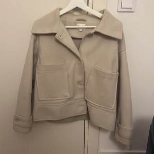 Beige ulljacka med stora fickor - Snygg beige jacka från Arket i ullmix med oversized krage och stora, fyrkantiga fickor framtill. Jackan har knäppning med knappar och breda ärmslut med knappdetalj. Perfekt för dig som gillar minimalistisk och clean stil.