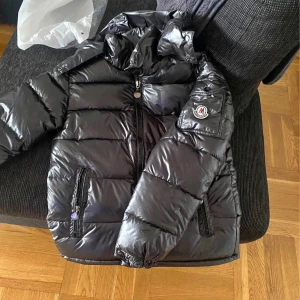 Svart pufferjacka från Moncler - Snygg svart pufferjacka från Moncler med glansig finish och klassisk logga på ärmen. Jackan har huva, dragkedjor på fickorna och framtill, samt quiltad design som håller värmen. Perfekt för kalla dagar och riktigt stilren look.