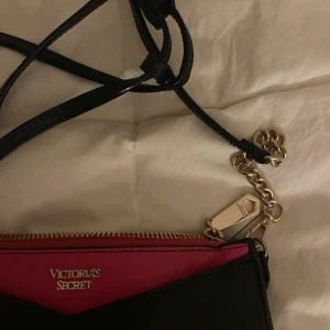 Svart axelväska - Snygg svart axelväska med detaljer i guld och en cool rosa insida. Väskan har en kedja och dragkedja upptill, samt en smal axelrem. Perfekt storlek för det viktigaste och har en stilren, modern look med exklusiv känsla.