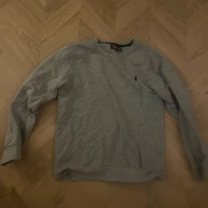 Grå sweatshirt från MCA köpt från Åhléns  - En stilren grå sweatshirt från Polo Ralph Lauren med klassisk rund hals och diskret broderad logga på bröstet. Tröjan är långärmad och har ribbade muddar vid ärmslut och nederkant. Perfekt för en chill och avslappnad look. 