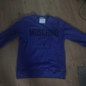Lila Moschino sweatshirt med tryck - Snygg lila sweatshirt från Moschino med stort svart logotryck på bröstet. Tröjan har rund halsringning och långa ärmar. Perfekt för dig som vill sticka ut med ett exklusivt streetwear-plagg från Milano.