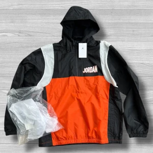 Jordan vindjacka  - Jordan vindjacka i svart, orange och vitt med huva och dragkedja. Stor logga på ryggen och JORDAN-tryck på bröstet. Jackan har meshfoder och elastiska muddar. Perfekt för dig som vill sticka ut med sportig streetstyle.