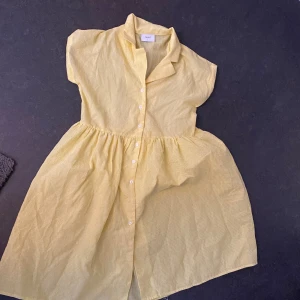 Gul kortklänning från Grunt - Söt gul kortklänning från Grunt med små vita knappar framtill, korta ärmar och krage. Klänningen har en lätt rynkad kjol och är tillverkad i ett tunt, luftigt bomullstyg – perfekt för varma dagar.