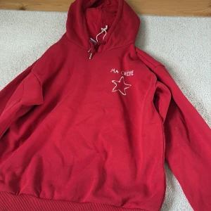 Röd hoodie med broderad stjärna - Röd hoodie med huva och snörning, broderad vit stjärna och texten 'MA CHÉRIE' på bröstet. Mjuk bomullskänsla och ribbade muddar. Perfekt för en chill och avslappnad stil.