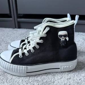 Karl Lagerfeld Converse  - Svarta höga sneakers från Karl Lagerfeld med vit plattformssula och vita detaljer i storlek 38. Skorna har snörning, rund tå och en cool Karl Lagerfeld-figur på sidan. Tillverkade i canvas med breda vita dragflikar baktill och logga på sulan. Perfekt för dig som gillar statement pieces.