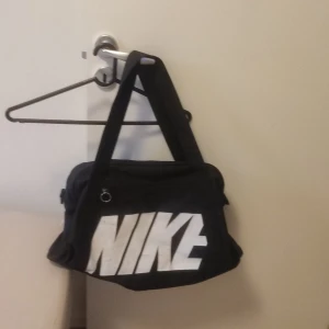 Svart träningsväska från Nike - Stor svart träningsväska från Nike med tydlig vit logga på framsidan. Väskan har dubbla handtag, dragkedja upptill och en rymlig insida. Perfekt för gymmet eller sportaktiviteter. Tillverkad i slitstark polyester.