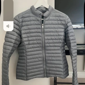 Grå dunjacka från Colmar Originals - Snygg grå dunjacka från Colmar Originals med quiltad design och dragkedja framtill. Jackan har en stilren, smal passform och är lätt vadderad, perfekt för kyliga dagar. Insidan är ljusgrön och jackan har ståkrage och långa ärmar. Jackan är i liten storlek. Och den är grå grön i färg 