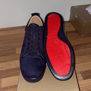 Mörkblå sneakers från Christian Louboutin - Exklusiva mörkblå sneakers från Christian Louboutin i mocka med ikonisk röd sula. Skorna har rund tå, platt sula och klassisk snörning. Insidan är i beige skinn med guldfärgad logga. Perfekt för dig som vill sticka ut med lyxig streetstyle.