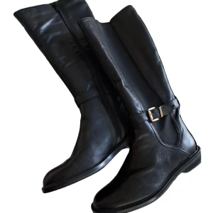 Svarta höga stövlar i skinn fr. Anna Field  - Snygga svarta stövlar med platt sula och rund tå, tillverkade i äkta skinn. De har en dekorativ rem med metallspänne och dragkedja på sidan för enkel på- och avtagning. Perfekta för dig som gillar stilrena och klassiska boots med lite edge.