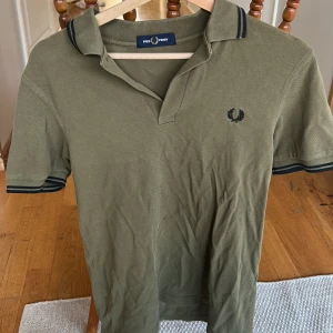 Olivgrön Fred Perry piké t-shirt - Snygg olivgrön piké från Fred Perry med klassisk krage och korta ärmar. T-shirten har marinblå detaljer på krage och ärmslut samt Fred Perry-loggan broderad på bröstet. Tillverkad i mjuk bomull som känns skön mot huden.