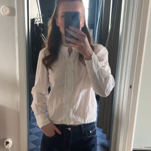 Vit blus från Zara - Vit blus från Zara med volangdetaljer längs knappraden och runt halsen. Blusen har lång ärm och rundad hals med knappar framtill. Använd fåtal gånger. 