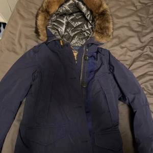Mörkblå Parajumpers vinterjacka med päls - Säljer en mörkblå Parajumpers vinterjacka med stor huva och pälsdetalj. Jackan har flera fickor, dragkedja och knappar framtill samt quiltat foder i silver. Perfekt för kalla dagar och riktigt snygg streetstil. Storlek ungefär 150cm (barn)
