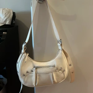 Vit axelväska från Monki i skinnimitation - Snygg vit axelväska från Monki i skinnimitation med silvriga nitar och dragkedjor. Väskan har justerbart axelband med metallringar.