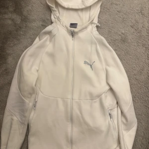 Vit hoodie med dragkedja från Puma - Snygg vit hoodie från Puma med hel dragkedja och huva. Tröjan har två fickor med dragkedja och diskret Puma-logga på bröstet. Perfekt för en sportig och clean look. Mjukt material och bekväm passform. Storlek M men sitter som S.