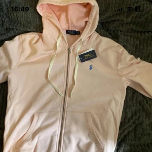 Ljusrosa hoodie från Polo Ralph Lauren - Säljer en ljusrosa hoodie från Polo Ralph Lauren med dragkedja och huva. Tröjan har vita snören, två fickor fram och det klassiska blå logobroderiet på bröstet. Perfekt för en chill och stilren look+ den är jätte skön.