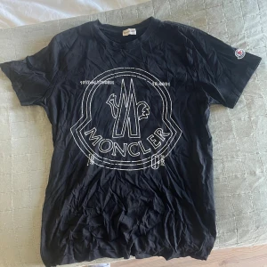 Svart Moncler t-shirt med stor logga - Svart t-shirt från Moncler med stor vit logga framtill och broderad patch på ärmen. Klassisk rund hals och korta ärmar. Tillverkad i mjuk bomull som känns skön mot huden. Perfekt för dig som gillar streetwear och vill sticka ut med en ikonisk design. QR kod finns 