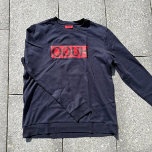 Svart långärmad tröja från Hugo boss  - Snygg svart långärmad tröja från Hugo boss med röd logga framtill. Tröjan har en klassisk rund halsringning och är gjord i mjuk bomull. Perfekt för dig som gillar stilrena plagg med tydlig branding.