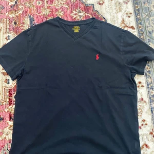 Svart t-shirt från Polo Ralph Lauren - Svart t-shirt från Polo Ralph Lauren med klassisk v-ringning och röd broderad logga på bröstet. Tillverkad i mjuk bomull och har en normal passform. Perfekt för en clean och stilren look.