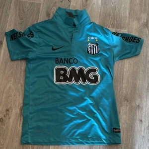 Santos Neymar Jr blå fotbollströja Nike - Säljer en blå Santos FC fotbollströja från Nike med Neymar Jr och nummer 11 på ryggen. Tröjan har korta ärmar, ståkrage och tryckta sponsorer som BMG och Netshoes. Materialet är lätt och andas, perfekt för träning eller match. Snygg design med klubbmärke och detaljer i svart och vitt.