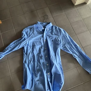Blå skjorta från Polo Ralph Lauren - Klassisk blå skjorta från Polo Ralph Lauren med button down-krage och broderad logga på bröstet. Skjortan har långa ärmar, vita knappar och är tillverkad i mjuk bomull. Perfekt för en clean och stilren look.