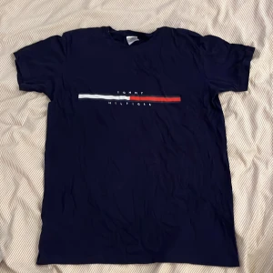 Marinblå t-shirt Tommy Hilfiger - Snygg marinblå t-shirt från Tommy Hilfiger med klassisk logga och tryck i vitt och rött på bröstet. T-shirten har rund hals och korta ärmar, perfekt för en chill och stilren look. Materialet är mjuk bomull som känns skönt mot huden.