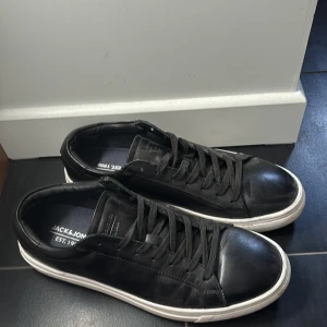 Svarta sneakers från Jack & Jones - Svarta sneakers från Jack & Jones med klassisk design och vita sulor. Skorna har rund tå, snörning och är tillverkade i skinnliknande material. Perfekta för en clean och stilren look. Storlek 42. Hjälper min brorsa att sälja dessa, de är lite dammiga men det går bara att borsta bort. Pris kan diskuteras vid snabbt köp 
