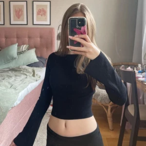 Svart ribbad croppad topp - Svart långärmad croppad topp med ribbad struktur och vid, öppen rygg. Toppen har en tight passform och vida ärmar som ger en snygg siluett. Perfekt att styla med höga jeans eller kjol för en trendig look.