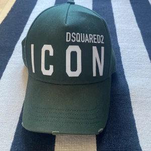 Grön Dsquared2 ICON keps - Mörkgrön keps från Dsquared2 med stor vit 'ICON'-text framtill och 'DSQUARED2' ovanför. Baksidan har broderad text 'DEAN & DAN CATEN'. Klassisk böjd skärm och justerbar passform. Tillverkad i bomull, perfekt för en streetwear-look.