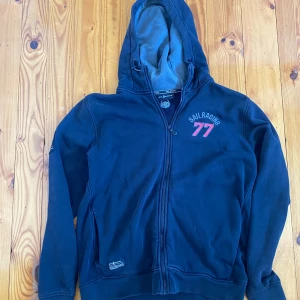 SAIL RACING HOODIE SVART - Svart hoodie från Sail Racing med dragkedja och huva. Snyggt tryck med 'Sail Racing 77' i rött och vitt på bröstet. Mjuk insida och praktiska fickor framtill. Perfekt för chill dagar eller när du vill ha en sportig look.