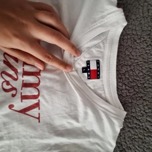 Vit t-shirt från Tommy Jeans - Vit t-shirt från Tommy Jeans med röd logga tryckt framtill. Klassisk rund halsringning och korta ärmar. Skön och enkel modell i bomull, perfekt till jeans eller shorts. Snygg och stilren basic med ikonisk branding. 