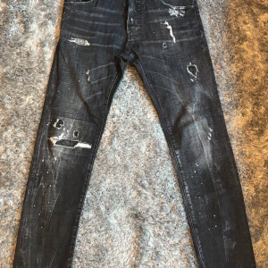 Svarta Dsquared2 jeans med slitningar - Snygga svarta jeans från Dsquared2 med coola slitningar, färgstänk och patchdetalj på bakfickan. Smal passform och klassisk femficksdesign. Jeansen har en unik streetkänsla och är tillverkade i bomull med stretch för extra komfort.
