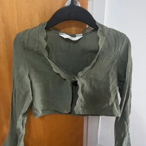 Olivgrön croppad blus från H&M - Säljer en olivgrön croppad blus från H&M med långa ärmar och v-ringning. Blusen har fina spetsdetaljer längs kanten och knyt framtill. Materialet är lätt och har en subtil struktur som ger en snygg look.