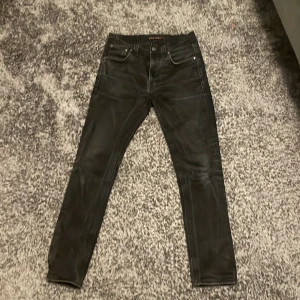 Svarta jeansbyxor från Nudie Jeans W 31 L 32 - Säljer ett par svarta jeansbyxor från Nudie Jeans med klassisk femficksdesign och smal passform. Byxorna har en stilren look med svarta sömmar och läderpatch med logga bak i midjan. Perfekta för dig som gillar en clean och modern stil.