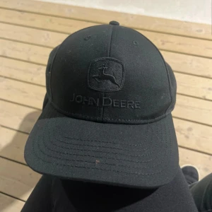 Svart John Deere keps med broderi - Säljer en svart keps från John Deere med snyggt broderat logotyp framtill. Klassisk böjd skärm och justerbar snapback baktill för perfekt passform. Kepsen är stilren och passar dig som gillar enkel design och vill ha ett coolt märke på huvudet.