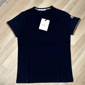 Mörkblå Mon*ler t-shirt med logga. Blå - Snygg mörkblå t-shirt från Mon*ler med klassisk rund hals och korta ärmar. Diskret Mon*ler-logga på vänster ärm och coola rödvita detaljer vid ärmslut och halsringning. Tillverkad i mjuk bomull som känns skön mot huden. Perfekt för dig som gillar stilrena märkesplagg.