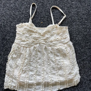 Vit volangig linnetopp med smala axelband - Supersöt vit linnetopp med smala axelband och volangdetaljer över hela toppen. Den har en lätt rynkad struktur och en feminin v-ringning. (Pris kan diskuteras)