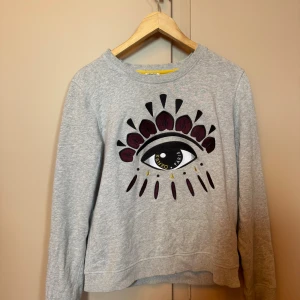 Grå Kenzo tröja med ögonmotiv - Nu säljer jag denna fina Grå sweatshirten från Kenzo med fint broderat ögonmotiv i svart, vinrött och gult på bröstet. Jag köpte den i storlek L på barnavdelningen och den passar mig som har storlek s i vuxenavdelningen. Jag säljer den för 300kr + frakt som kunden betalar, pris kan diskuteras.