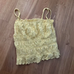 Gul broderad linnetopp från BikBok - Supersöt ljusgul linnetopp från BikBok med smala axelband och broderade detaljer. Toppen är smockad och har volangkant nertill, perfekt för sommarens alla tillfällen. Luftig och lätt i materialet, passar till både jeans och kjol.