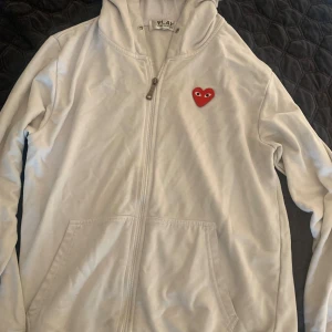 Vit hoodie från Comme des Garçons Play - Vit hoodie från Comme des Garçons Play med dragkedja och huva. Tröjan har det ikoniska röda hjärtat med ögon broderat på bröstet och två fickor framtill. Tillverkad i mjuk bomull, perfekt för en chill och stilren look.